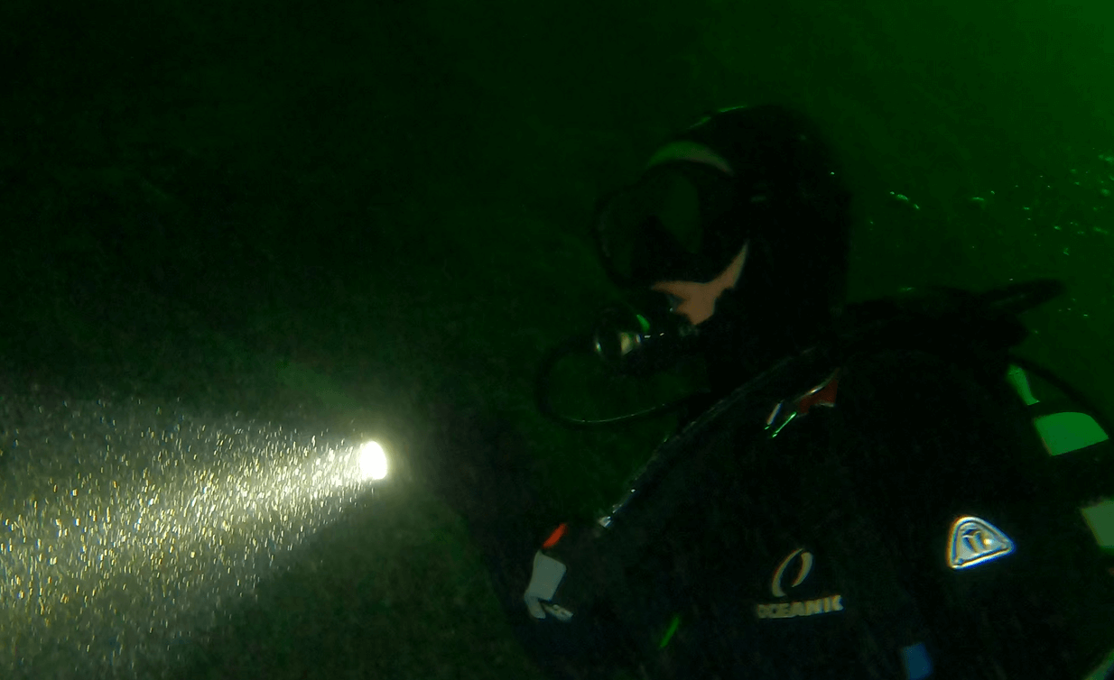 Taucher leuchtet mit Tauchlampe und trägt eine Kopfhaube in dunklem Wasser