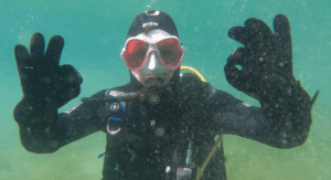Taucher schaut mit leerer Tauchmaske frontal in die Kameras und gibt mit beiden Händen das OK Zeichen
