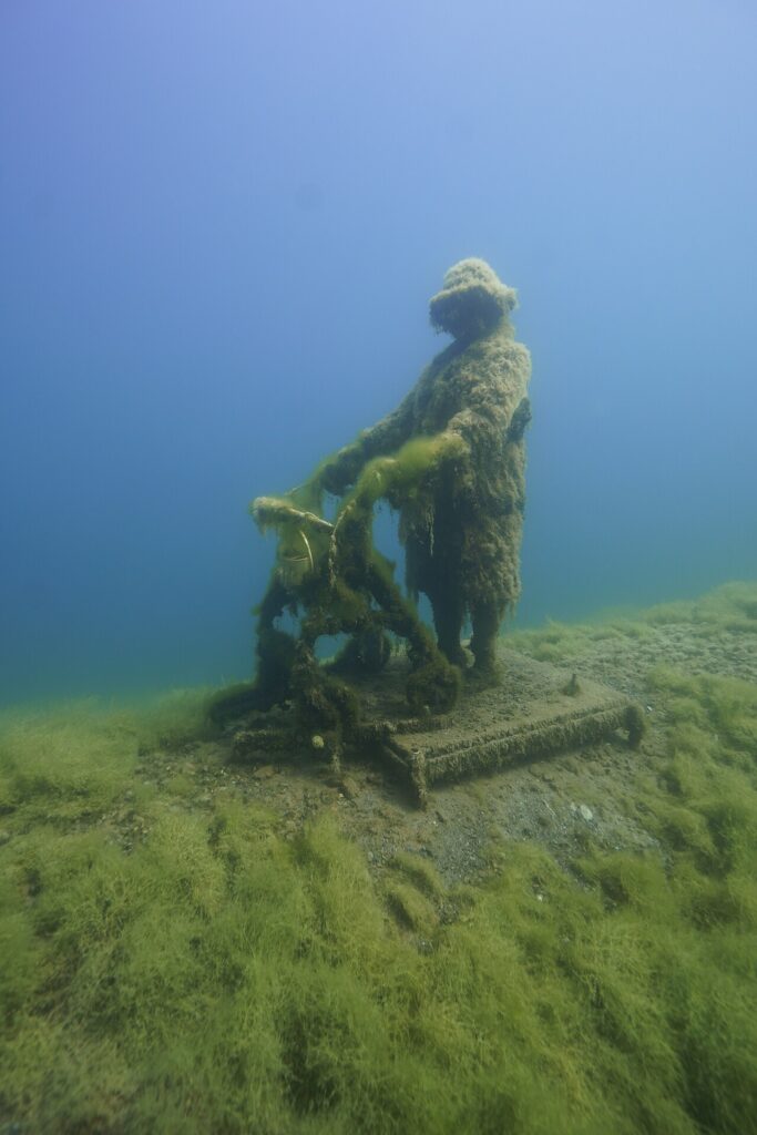 Mit Asorry lgen bewachsene Betonfigur im Sundhäuser See